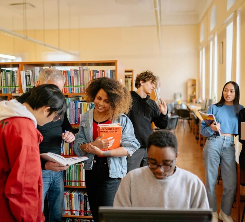 A diverse group of students studying together at a university library, engaging with books and technology.